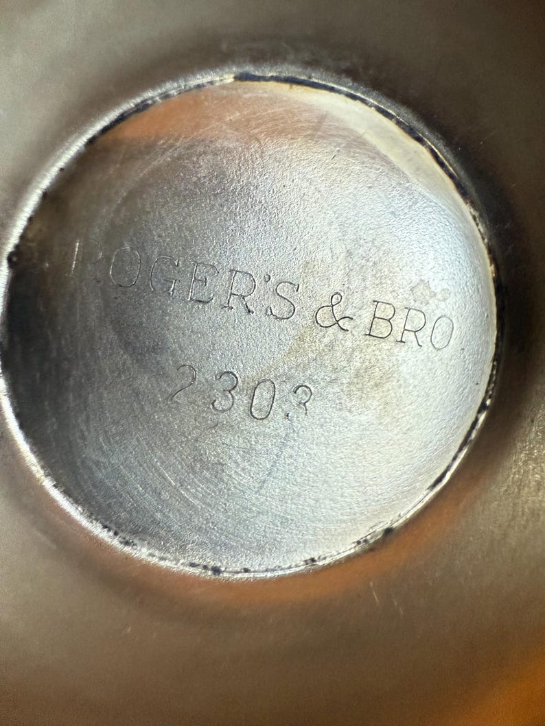 May include: Close-up of a silver-colored metal object with the text "ROGERS & BRO" and the number "2303" etched into the surface. The object has a circular shape with a slightly textured appearance.