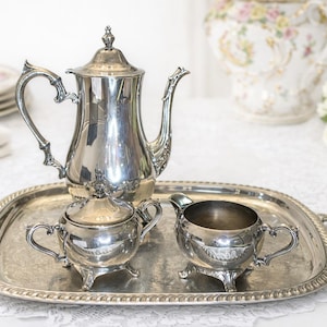 Puede incluir: Un juego de té plateado sobre una bandeja plateada. El juego incluye una tetera, un azucarero y una lechera. La bandeja tiene asas y un borde decorativo. El juego se exhibe sobre un mantel de encaje blanco.
