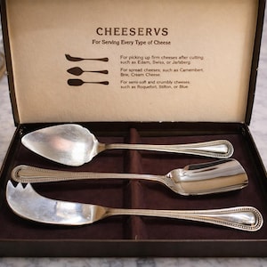 May include: A set of three silver cheese serving utensils in a brown box. The box lid reads "CHEESERVS For Serving Every Type of Cheese." The utensils include a cheese knife, a scoop, and a server.