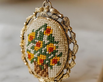 May include: Vintage gold-tone pendant necklace featuring a cross-stitch floral design. The oval pendant showcases a detailed needlepoint of yellow and red flowers in a brown pot, framed by an ornate gold border and chain.