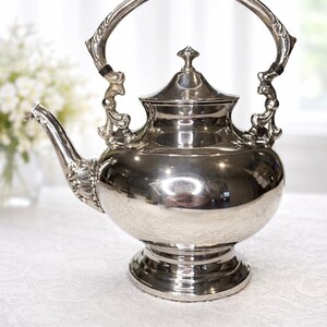May include: A silver-colored teapot with a rounded body, a curved spout, and a large, arched handle. The teapot has a decorative lid and sits on a pedestal base. The design includes ornate detailing.