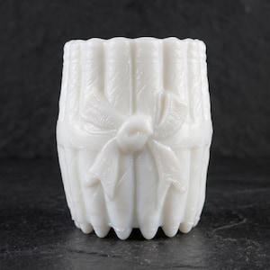 May include: A white milk glass vessel featuring a vertical ribbed texture and a molded bow detail on the front. The container has a scalloped top edge and a scalloped base, with a semi-translucent finish that catches the light across its surface.