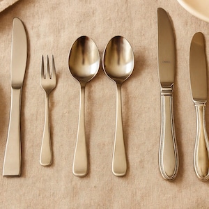 May include: A set of stainless steel silverware, including knives, forks, and spoons, arranged on a tan linen tablecloth. The silverware has a simple, elegant design, with a matte finish. A white plate is visible in the background.