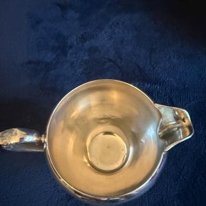 May include: A shiny, silver-colored creamer with a handle and a spout. The creamer is round with a smooth, reflective surface. The creamer is set against a dark blue background.