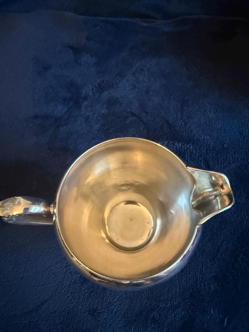 May include: A shiny, silver-colored creamer with a handle and a spout. The creamer is round with a smooth, reflective surface. The creamer is set against a dark blue background.