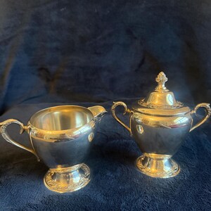 May include: A silver creamer and covered sugar bowl set. The creamer has a curved handle and spout. The sugar bowl has two handles and a decorative lid. Both pieces have ornate detailing around the base and are set against a dark blue background.