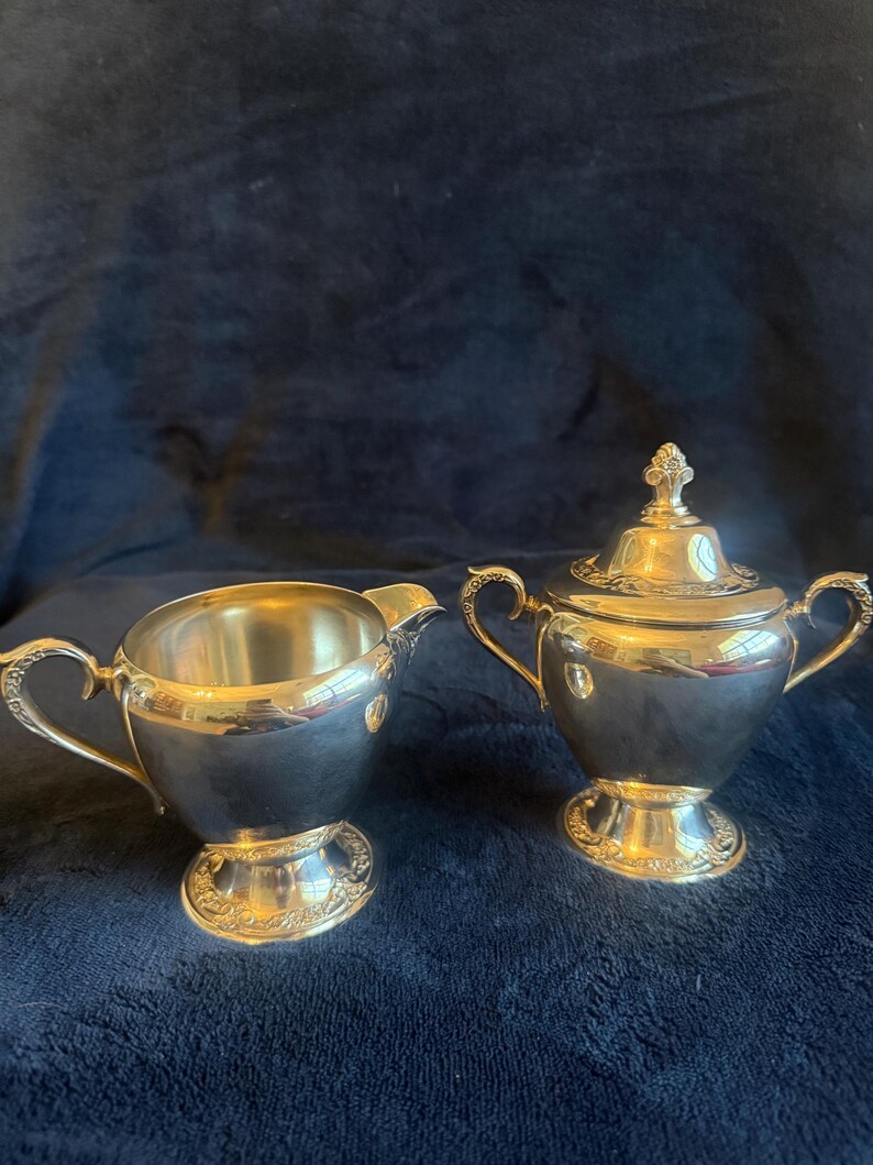 May include: A silver creamer and covered sugar bowl set. The creamer has a curved handle and spout. The sugar bowl has two handles and a decorative lid. Both pieces have ornate detailing around the base and are set against a dark blue background.