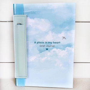 Peut inclure: Un journal avec une couverture bleu clair représentant un ciel nuageux. Le texte "A place in my heart Grief Journal" est imprimé sur la couverture. Un porte-stylo bleu clair est attaché au journal avec un élastique bleu.