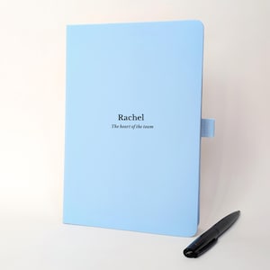 May include: A light blue notebook with a fabric pen loop on the side. The cover features the printed text "Rachel" in a bold font above the smaller text "The heart of the team". A black ballpoint pen is positioned at the bottom right corner.
