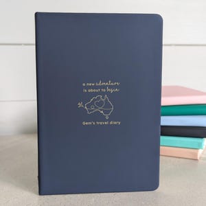 May include: A dark blue travel journal with a gold-coloured illustration of Australia and the text "a new adventure is about to begin". The words "Gem's travel diary" are printed below. Other coloured journals are stacked in the background.