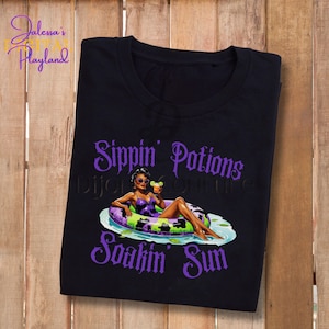 Op de afbeelding: Zwart T-shirt met een paarse afbeelding van een vrouw in badpak op een opblaasbare ring, met een cocktail. De tekst "Sippin' Potions Soakin' Sun" staat in paarse letters. Het shirt heeft een ronde hals.
