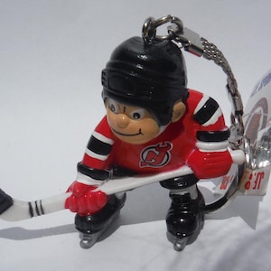 May include: A red, white, and black hockey player keychain with a stick and a New Jersey Devils logo. The keychain is attached to a silver chain.