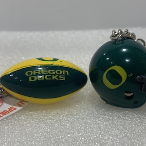 May include: Two keychains featuring the Oregon Ducks logo. One keychain is a green and yellow football, the other is a green football helmet with a black facemask.