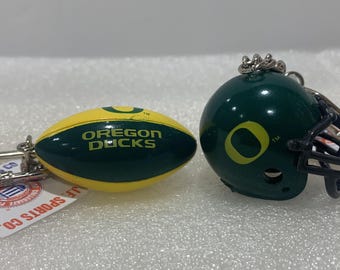 May include: Two keychains featuring the Oregon Ducks logo. One keychain is a green and yellow football, the other is a green football helmet with a black facemask.