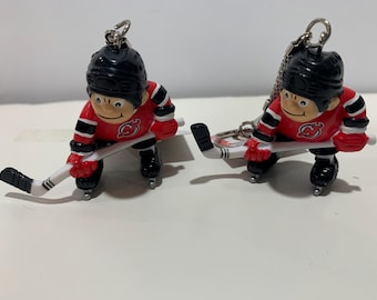 May include: Two red and black hockey player keychains with white sticks and a New Jersey Devils logo. The keychains are on a white surface.