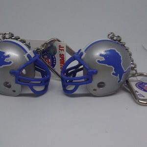 May include: Two silver football helmet keychains with blue and white Detroit Lions logos. The keychains are attached to silver key rings with a white tag that reads "J.F. Sports Co."