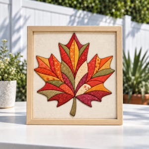 May include: A square framed wall art piece featuring a central maple leaf design composed of various fabric segments in shades of red, orange, yellow, and olive green. The leaf is set against a plain off-white background within a light wood frame.