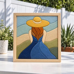 May include: A square framed wall art piece featuring a layered fabric collage of a person seen from behind. The figure wears a wide-brimmed yellow hat and a flowing blue dress, set against a background of abstract rolling hills in shades of green and tan.