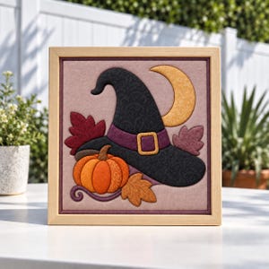 May include: A square framed fabric wall art piece featuring a black witch hat with a purple band and gold buckle, a bright orange pumpkin, a yellow crescent moon, and several autumn leaves in shades of red, brown, and gold on a mauve background.