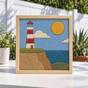 May include: A square framed fabric wall art piece featuring a lighthouse with red and white stripes on a cliffside. The scene includes a yellow sun, white clouds, a blue sky, a dark blue ocean, and green grassy areas, all rendered in layered fabric.