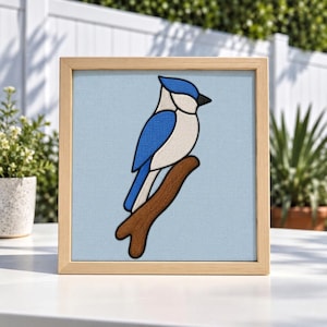 May include: A square framed wall art piece featuring a stylised blue jay illustration. The bird has a bright blue wing and tail with a white chest and black beak, perched on a brown branch against a solid light blue background in a wood frame.