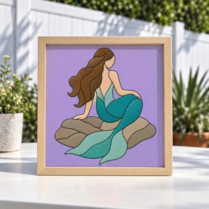May include: A square framed wall art piece featuring a stylized illustration of a mermaid with long brown hair, sitting on a tan rock. The mermaid has a teal and light blue tail, set against a solid light purple background in a light wood frame.