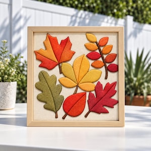 Puede incluir: Una pieza de arte mural cuadrada con marco de madera clara y fondo de tela color crema. La obra muestra seis hojas de fieltro distintas en tonos rojo, naranja, amarillo y verde oliva, dispuestas en un grupo para representar el follaje otoñal.