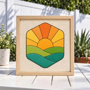 May include: A square wooden frame containing a centered hexagonal artwork. The design features a stylized orange and yellow sun rising over layered green and teal rolling hills, all outlined in black against a light beige fabric background.