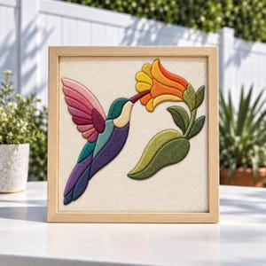 Puede incluir: Un cuadro cuadrado con una aplicación de tela que muestra un colibrí en tonos rosa, turquesa y morado, volando cerca de una gran flor naranja y amarilla con hojas verdes sobre un fondo de tela de color blanco roto.