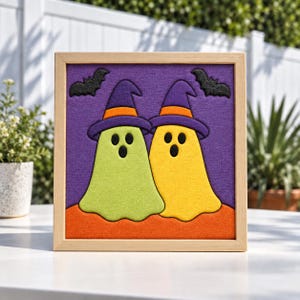 May include: A square framed wall art piece featuring a purple fabric background with two ghosts wearing purple and orange witch hats. One ghost is light green and the other is yellow. Two black bats are positioned above the ghosts on an orange base.