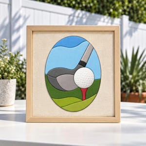 Op de afbeelding: Een vierkante houten lijst met een ovaalvormig gelaagd kunstwerk. Het kunstwerk toont een grijze golfclubkop achter een witte, getextureerde golfbal op een rode tee, tegen een achtergrond van groen gras en een blauwe lucht.