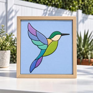 Puede incluir: Una pieza de arte mural cuadrada y enmarcada que presenta una ilustración estilizada de un colibrí sobre un fondo azul claro. El ave está compuesta por formas segmentadas en tonos verde, azul, morado y crema, delineadas con un borde negro.