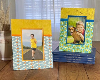 May include: Two decorative picture frames with photos, one featuring a boy running and the other a boy with a dog. The frames have yellow, blue, and orange patterns. The frames are on a wooden surface.