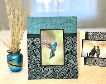 May include: A teal and black picture frame with a photo of a hummingbird. The frame has a textured teal top and a black and white swirl pattern. A second frame with a black and white swirl pattern is also visible.