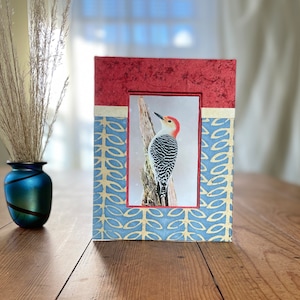 May include: A rectangular picture frame with a red top and blue patterned sides. The frame holds a picture of a red-bellied woodpecker perched on a tree branch. A blue vase with dried grass is on the left.