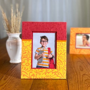 May include: A rectangular photo frame with a red top and a yellow patterned bottom. The frame holds a photo of a child wearing glasses, holding a tablet, and a red backpack. The frame is on a wooden surface.