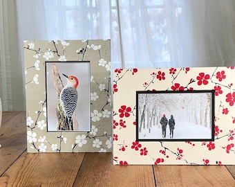 May include: Two decorative picture frames with floral patterns. One frame has a beige background with white flowers, the other has a beige background with red flowers. Both frames have black borders and display photographs.