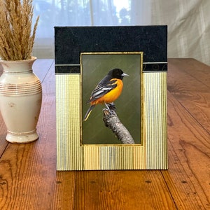 May include: A framed print of an orange and black bird perched on a branch. The frame has a black top and gold vertical striped sides. A cream-coloured vase with dried grass is to the left of the frame.