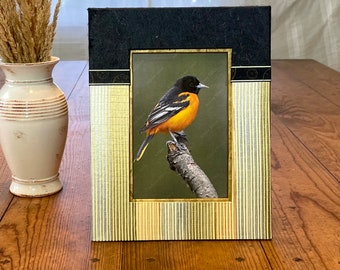 May include: A framed print of an orange and black bird perched on a branch. The frame has a black top and gold vertical striped sides. A cream-colored vase with dried grass is to the left of the frame.