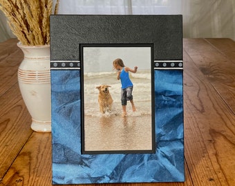 May include: A rectangular photo frame with a black top and blue textured paper bottom. The frame holds a photo of a child and a dog playing in the ocean. The frame has a black border around the photo.