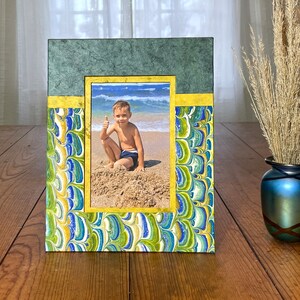 May include: A decorative picture frame with a photo of a child at the beach. The frame has a green top, gold accents, and a marbled pattern in blue, green, and yellow. The photo is framed in gold.