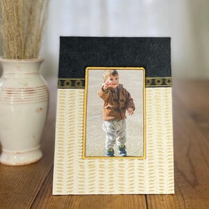 May include: A rectangular photo frame with a gold inner frame. The top section is black, and the bottom section is cream-coloured with a gold leaf pattern. The frame holds a photo of a young child wearing a brown jacket.