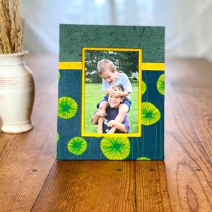 May include: A rectangular photo frame with a dark teal background and yellow floral pattern. The frame has a yellow border around the photo and a dark green top. The photo shows two children.