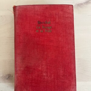 Pode incluir: Um livro vintage com capa vermelha. O título "Le Rouge et le Noir" está impresso em letras douradas. A capa do livro mostra sinais de desgaste, sugerindo sua idade. É um romance clássico.