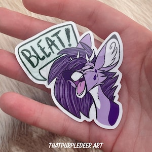 Könnte beinhalten: Lila und weißer Cartoon-Aufkleber mit einem pelzigen Wesen mit einer Sprechblase, auf der "BLEAT!" steht. Der Aufkleber befindet sich auf einer Hand.