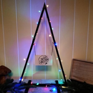 Może przedstawiać: Czarna drewniana rama w kształcie trójkąta z lampkami choinkowymi w kolorze czerwonym, niebieskim i zielonym. Rama ozdobiona jest tabliczką z napisem "Wesołych Świąt".