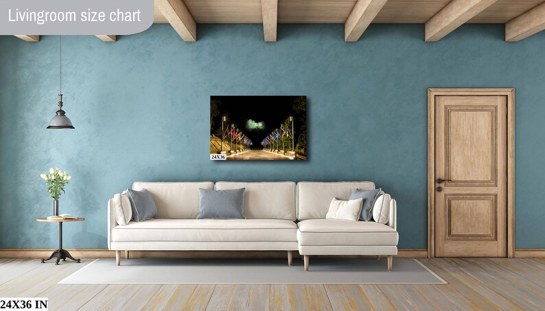 May include: A white couch with blue pillows sits in a living room with a blue wall and a wooden door. A framed photograph of a street lined with colorful flags hangs on the wall above the couch. The image is labeled "Livingroom size chart" and "24x36 in".