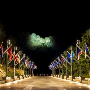 May include: A night view of Mount Rushmore National Memorial illuminated with spotlights. A walkway lined with flagpoles displaying various flags leads to the monument.