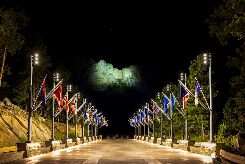 May include: A night view of Mount Rushmore National Memorial illuminated with spotlights. A walkway lined with flagpoles displaying various flags leads to the monument.