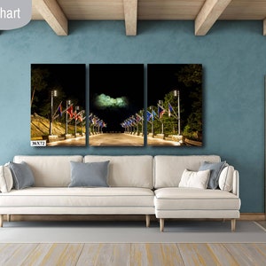 May include: A three-panel canvas print depicting a night scene of a street lined with flags and streetlights. The print is displayed on a white sectional sofa in a living room with a light blue wall and wooden beams. The text "36X72 IN" is visible in the bottom left corner of the image.